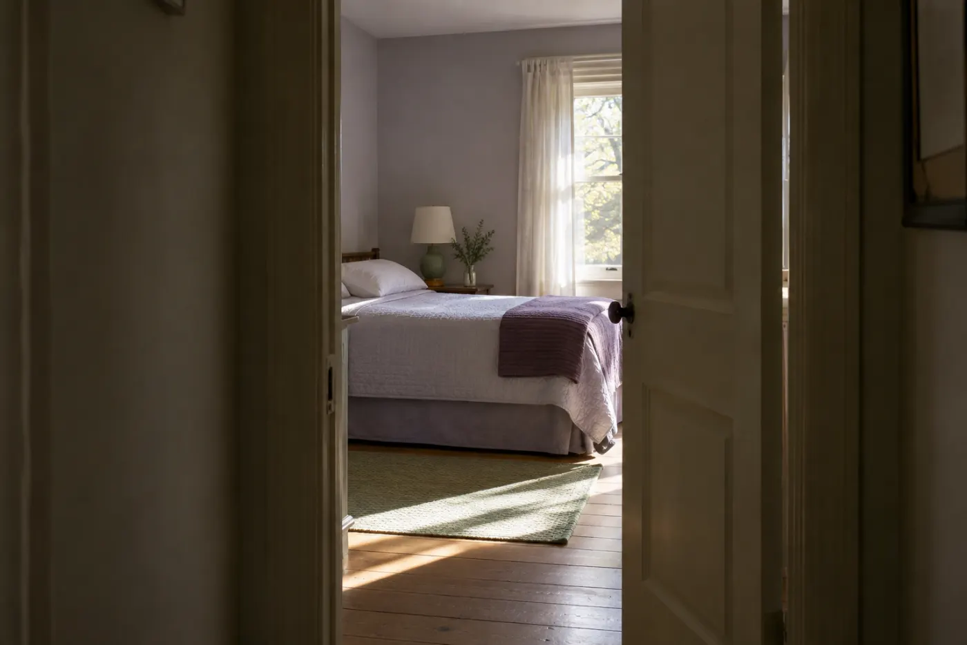 A hallway doorway half open onto a bright, plain bedroom: a made single bed with a folded blanket, a small lamp, a window with a sheer curtain and light on the floorboards.